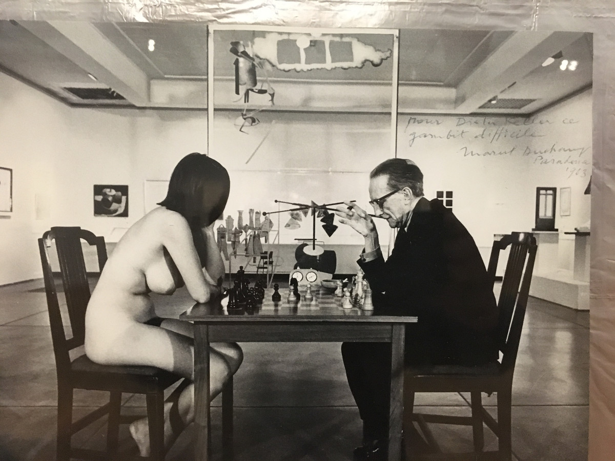 Duchamp war nicht nur ein Meister im Spiel mit der Kunst und der Selbstdarstellung, sondern auch ein Meister im Schachspiel. Das Foto von Julian Wasser zeigt ihn im Spiel gegen die 20jährige Künstlerin und Party-Gängerin Eve Babitz in der Duchamp-Retrospektive im Pasadena Art Museum 1963. Im Hintergrund das „Grosse Glas“.
