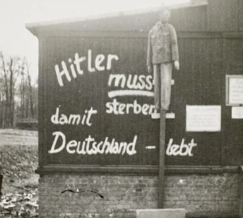 KZ Buchenwald im April 1945, Graffito auf einer Holzbaracke: „Hitler muss sterben damit Deutschland lebt“; davor eine aufgehängte Hitler-Puppe (Deutsches Bundesarchiv Bild 146-1978-134-24) 
